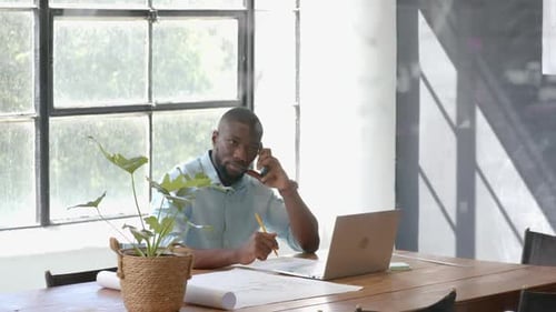 Talking on smartphone, man working on laptop and taking notes in office