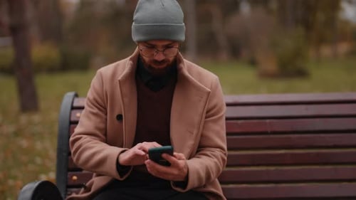 Young Man with Beard and Beanie Sitting on a Park Bench, Engaged with his Smartphone