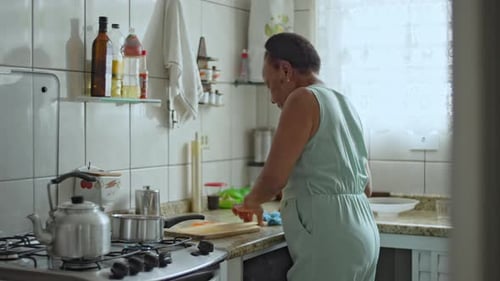 Elderly Woman Cleaning Food Waste From Cutting Board In Well Lit Kitchen With White Tiles