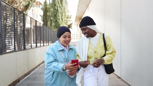 Zwei muslimische Freundinnen gehen spazieren, während sie ihr Handy benutzen, um lustige Inhalte in sozialen Medien zu teilen