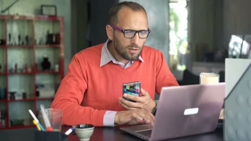 Man Uses Laptop and Phone at Table Indoors