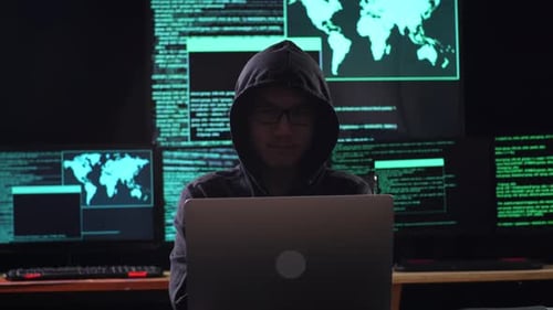 Hacker mit Kapuze schaut in die Kamera und verschränkt seine Arme, während er an einem Laptop arbeitet