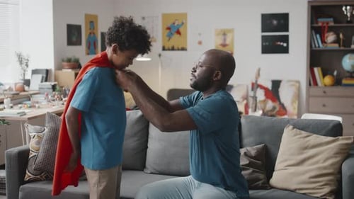 Black Dad Putting Cape on Little Son Playing Superhero at Home