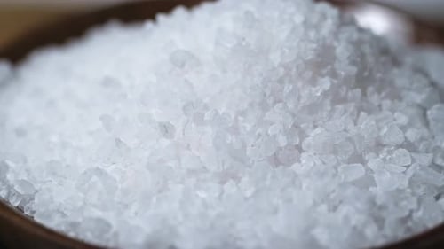 A close-up shot of coarse sea salt crystals in a wooden bowl. The white, irregularly shaped salt gra