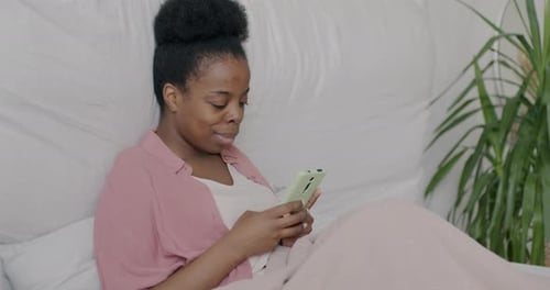 Cheerful African American Lady Using Smartphone Messaging Sitting in Bed in Apartment