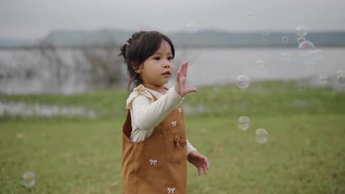 slow motion of happy toddler girl running and playing soap bubble in grass field near the river