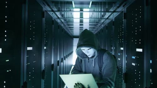 Mittelaufnahme eines maskierten Hackers in einem Hoodie, der mitten in einem Rechenzentrum voller Rack-Server steht