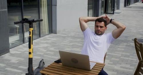 Man Sits in Street Cafe Working on Laptop Having Break with Hands Behind Head
