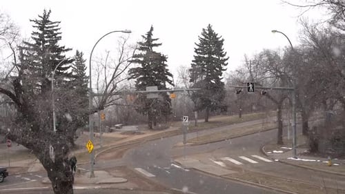 Intersection with some vehicles passing and very few people walking on the sidewalk with ligh snow f