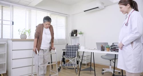 Asian senior woman walking slowly with walker during physical therapy in clinic while Caucasian