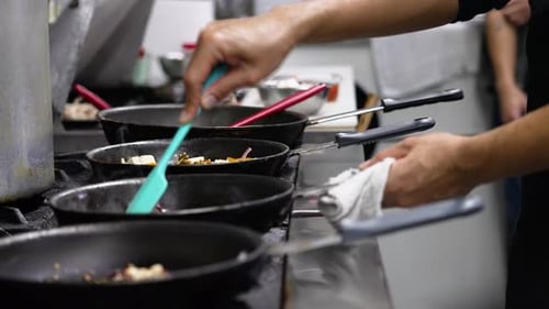 Restaurant cook stirs pan of food on stovetop alongside multiple skillets of cooking food, slider sl
