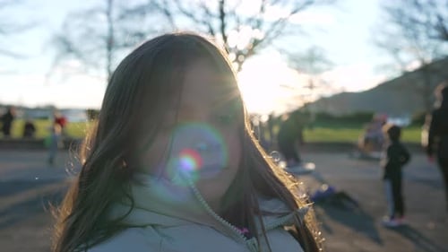 One pensive little girl standing at playground park during sunset and blurred people in background.