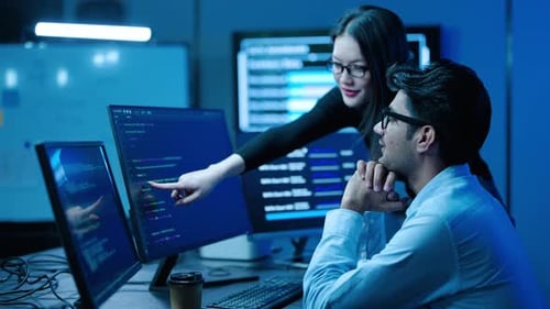 Two software engineers collaborate on computer coding projects in a high-tech office at night.