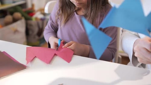 Young Child Focusing on Cutting Folded Pink Paper with Blue Handle Scissors at a White Table