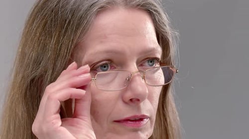 Blue Eyed Elderly Woman with Loose Hair Adjusts Elegant Glasses on Grey Background Closeup