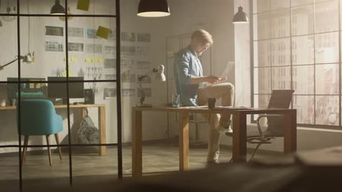 Creative Designer Walks around His Studio, Sits on His Desk Holds Laptop on the Knees while Working