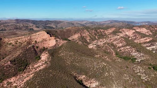 Drone footage flying above a rugged, hilly landscape with layered terrain, sparse vegetation, and a