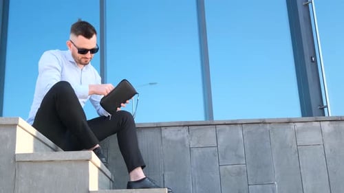 A Young Man Sits on the Steps in Front of the Office and Counts Money in His Wallet