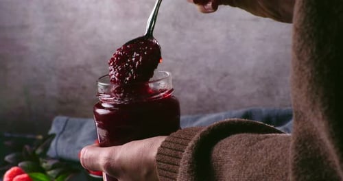 A close-up handheld shot of a jar of rich strawberry or raspberry jam being held while a spoon lifts