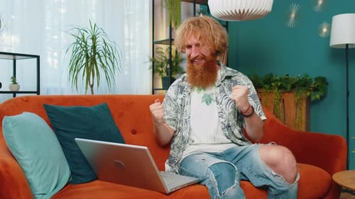 Red-Haired Man Uses Laptop and Cheers on Couch