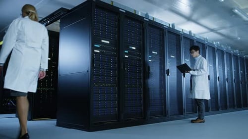 In Data Center: Male IT Specialist Wearing White Coat Walks Alongside Row of Server Racks, Uses Lap