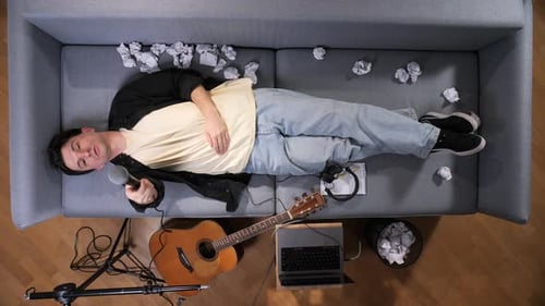 Young Man Holding a Microphone and Brainstorming Ideas on a Living Room Sofa Surrounded By Crumpled