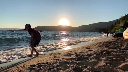 Garçon remplissant un seau de plage jaune avec de l'eau provenant du rivage accidenté. Soleil couchant sous la montagne