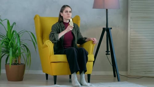 Woman Eating Banana Sitting in a Yellow Armchair