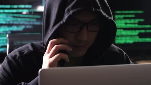 De perto de um hacker asiático com capuz falando no smartphone enquanto trabalha em um laptop