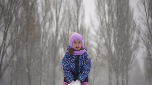 A Little Girl Stands Under Snowfall in the Park and Throws Snow A Girl in a Jacket and Hat Plays