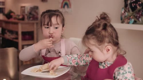 Girls eating pancake from plate at table, mother in background