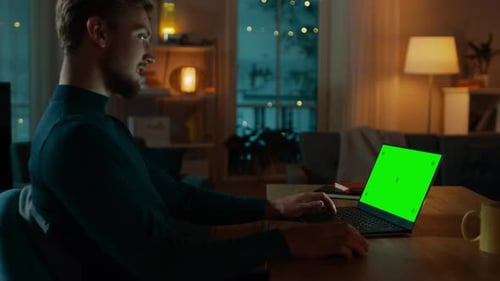 Man Sitting at His Desk Works on a Laptop with Green Mock-up Screen. Late at Night in His Living Ro