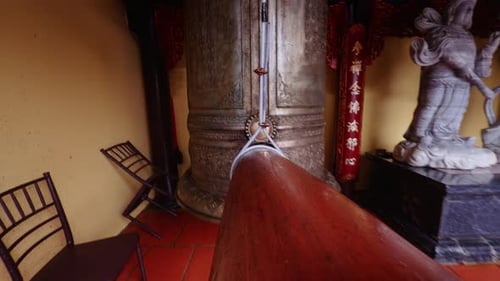 Longo batente de madeira que se estende em direção a um grande sino de bronze no interior do templo com estátuas ao fundo