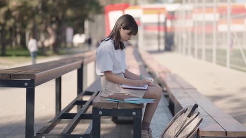 Schoolgirl Sits On Bench After Class and Writes In Notebook On Sports Field Near School