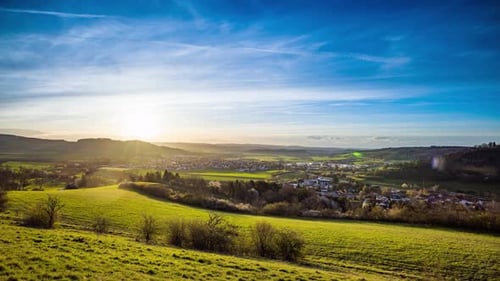 Sonnenuntergang beleuchtet eine Stadt in einem hügeligen Tal mit Wäldern und Wiesen