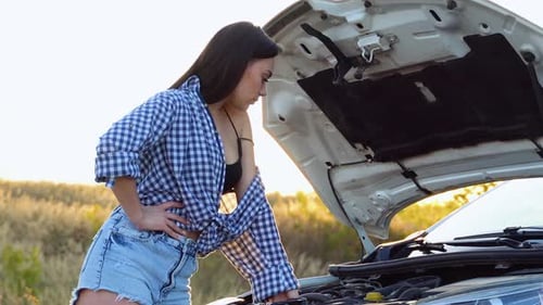 Beautiful Woman in Shirt and Shorts Looks in Open Car Hood on a Road