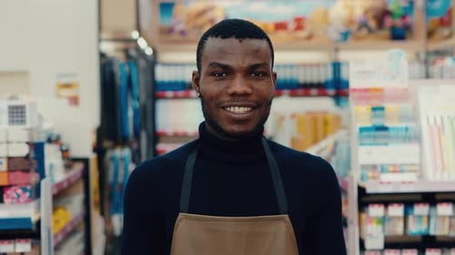 Smiling Male Store Clerk Wearing an Apron Standing in a Wellorganized Retail Store Aisle