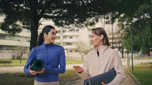 Two Women Walk and Talk in Park with Yoga Mats