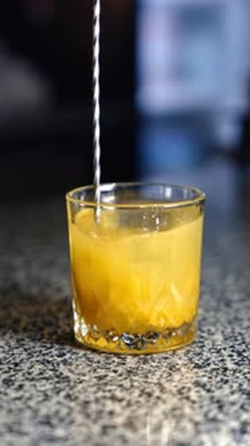 Yellow drink is mixed in the glass with an ice cube. Long spoon stirring the cocktail. Close up.