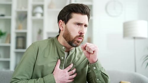 Hombre guapo tosiendo sentado en el sofá de la sala de estar de su casa. Enfermo de resfriado o gripe barbudo