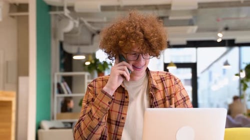 Young Freelancer or Blogger in Glasses with Braces Sitting in a Cafe Talking on Mobile Phone While