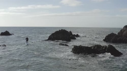 Aerial: Rough ocean water challenges stand up paddleboarder near rocks