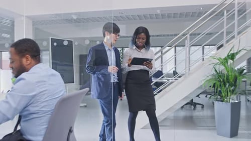 Two Young Male and Female Colleagues Walk in the Office Holding a Tablet Computer and Discuss Work.