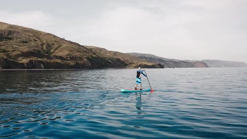 Professional Surfer Stands on Blue Board and Rows at Hills