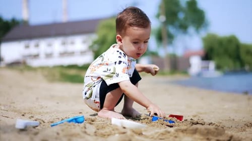 Cute Caucasian toddler sitting squatted on the beach. Kid takes some sand and throws it.