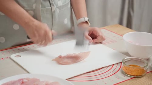 A cook is beating raw chicken breast with a meat tenderizer on a cutting board
