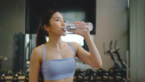 Portrait of tired asian woman in sportswear drinking water from bottle in gym