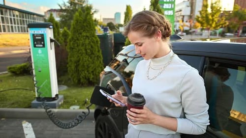 A young blonde woman with smartphone and coffee at a car charging station with charging electric car