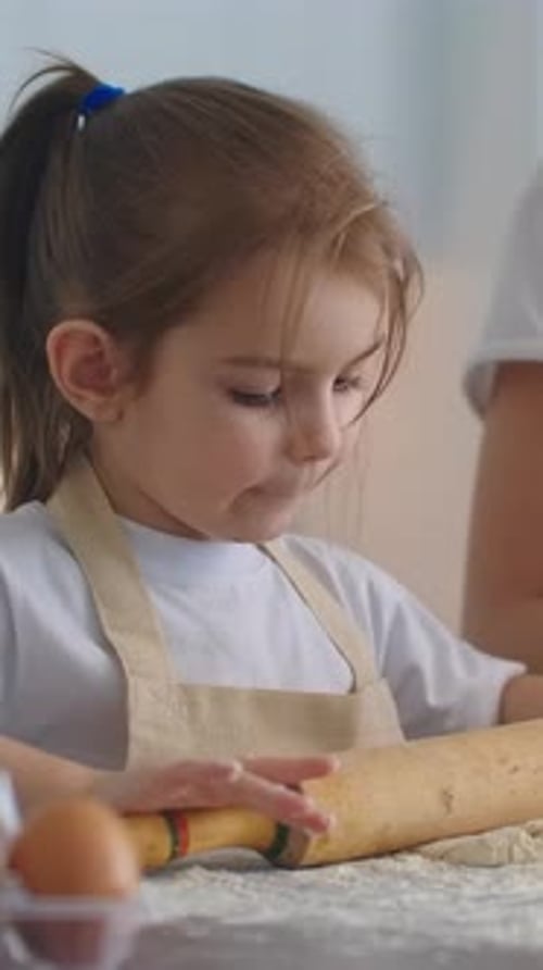 Little Girl in Apron Rolling Dough with Wooden Rolling Pin in Bright Kitchen Concentrating on Baking