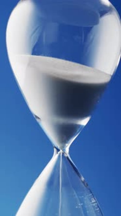 Vertical video of close up of hourglass with white sand pouring with copy space on blue background